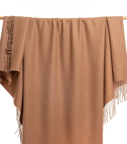 Prekrivac od Baby Alpake u camel boji prikazan na drvenoj šipki sa resama